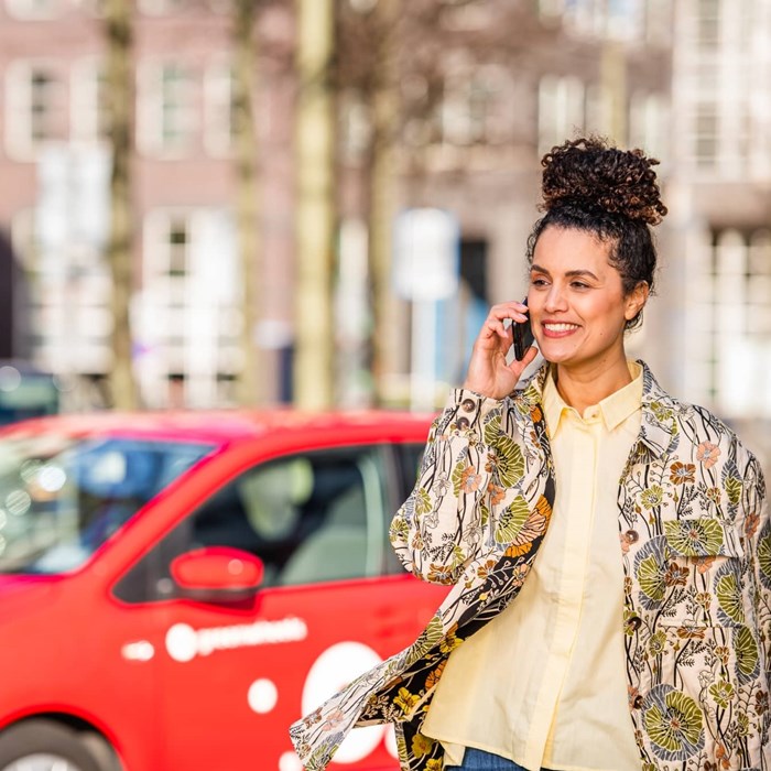 Vrouw met blazer en gele blouse en mobiele telefoon bij rode Greenwheels auto