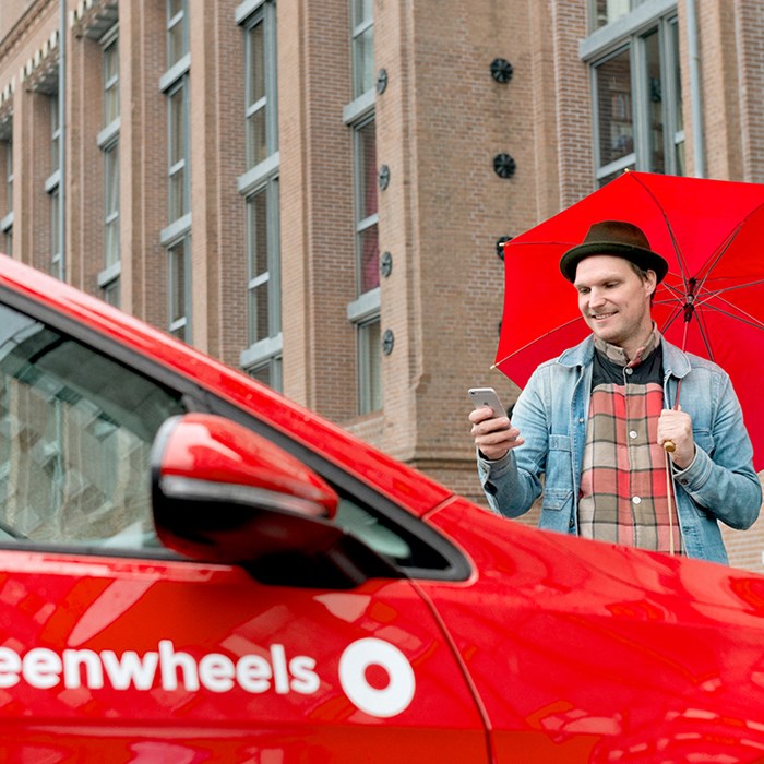 Man in regen op telefoon naast rode Greenwheels deelauto